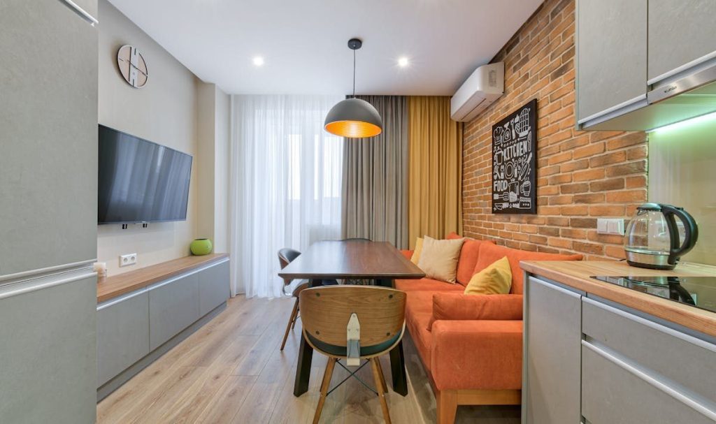 Studio com sofá laranja, mesa de refeições e cozinha junto à parede de tijolos aparentes